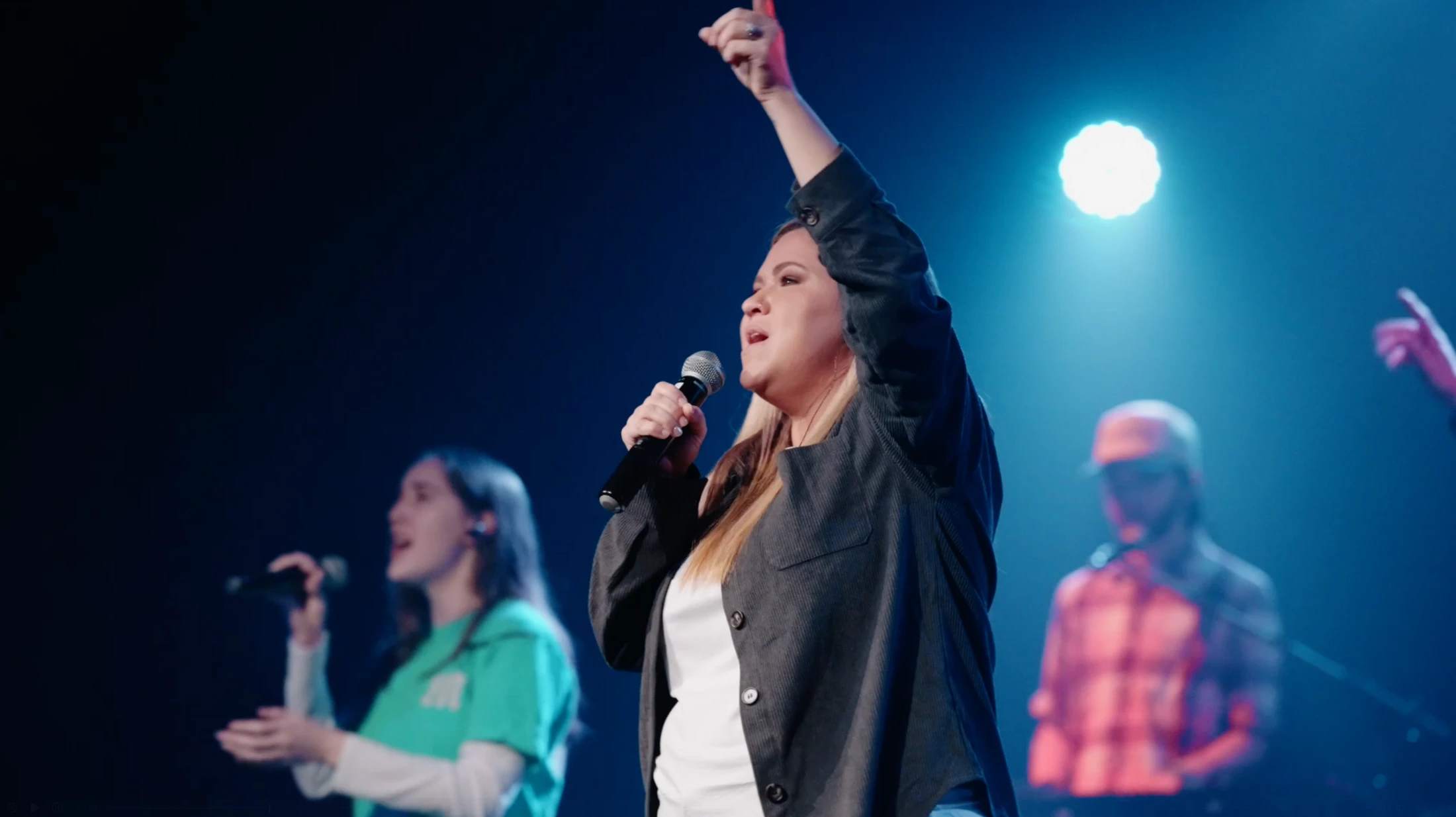 A woman with blonde hair sings into a microphone on a dimly lit stage, her arm raised. Other singers and musicians are blurred in the background.